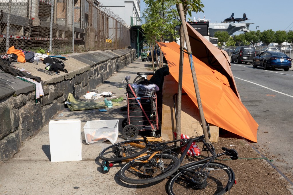 A homeless encampment with an orange tent, bikes, and scattered belongings on a sidewalk next to a street, with the Intrepid Museum and a spy plane in the background.