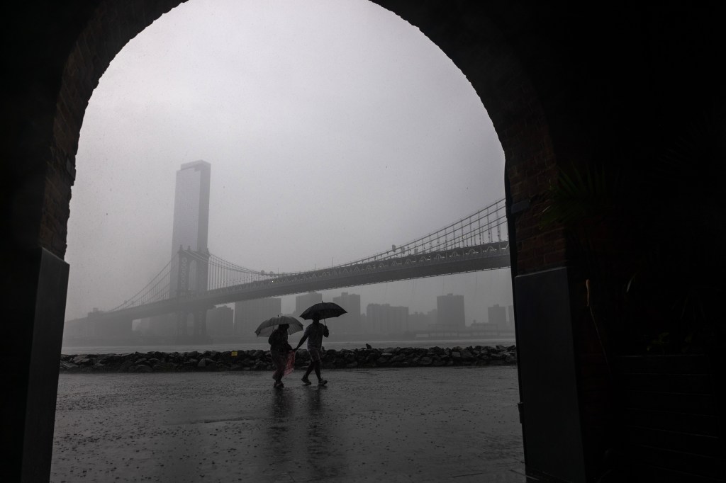Two people with umbrellas walk in the rain near the Manhattan Bridge and a tall building in Brooklyn.