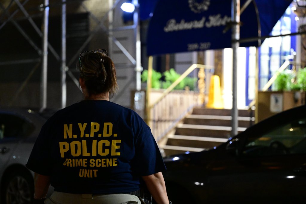 A NYPD Crime Scene Unit member stands with their back to the camera, facing the Belnord Hotel entrance.