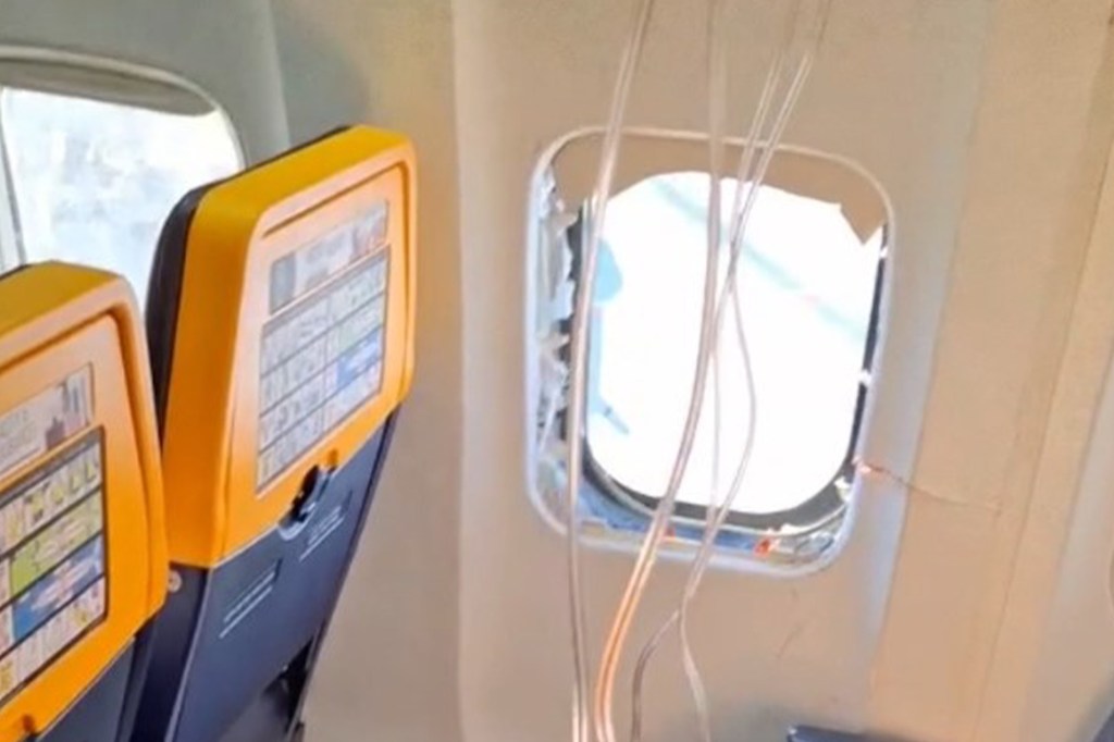 The interior of an airplane cabin shows a broken window with wires exposed.