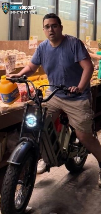 An individual with dark hair and glasses sitting on an electric bicycle, holding a jug of Sunny D, in what appears to be a market.
