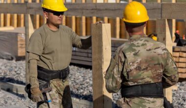 Two men wearing camouflage military uniforms and hard hats stand outside holding power tools next to a wooden building frame.