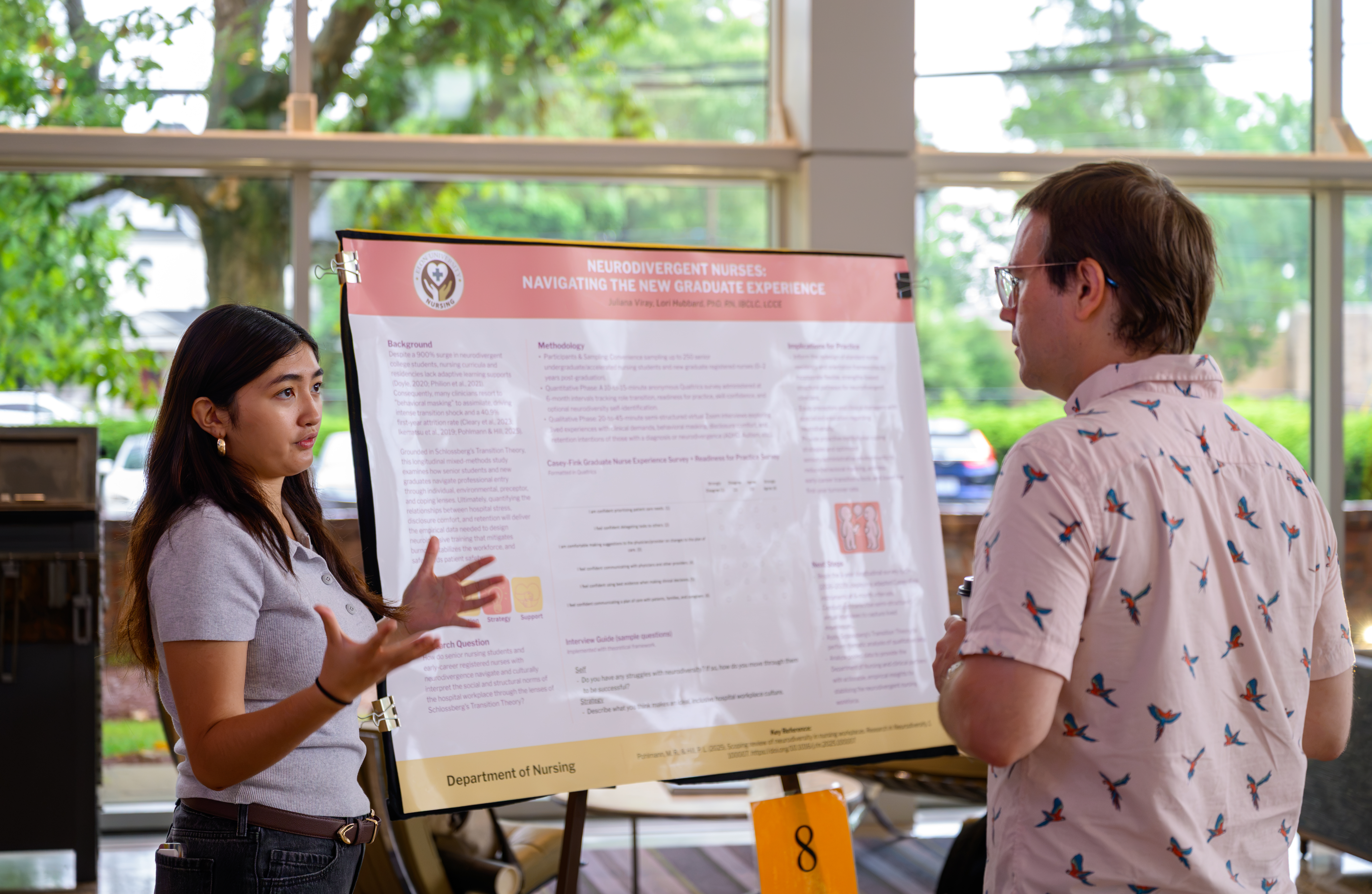 A young women presents a research presentation poster as a man listens and faces the poster