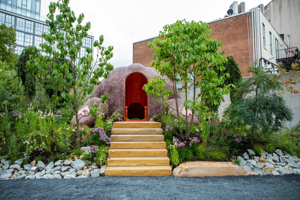 A unique outdoor structure surrounded by greenery and flowers, featuring steps leading to a red entrance.