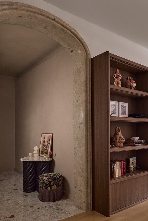 A cozy living space with a wooden bookshelf, decorative items, a side table, and artwork under a stone archway.