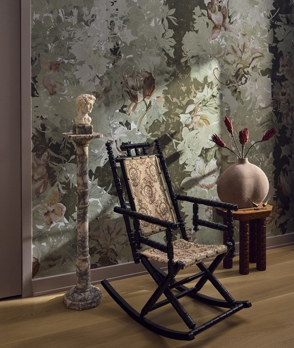 Vintage rocking chair next to a marble pedestal with a small statue, floral wallpaper, and a decorative vase on a wooden stool.