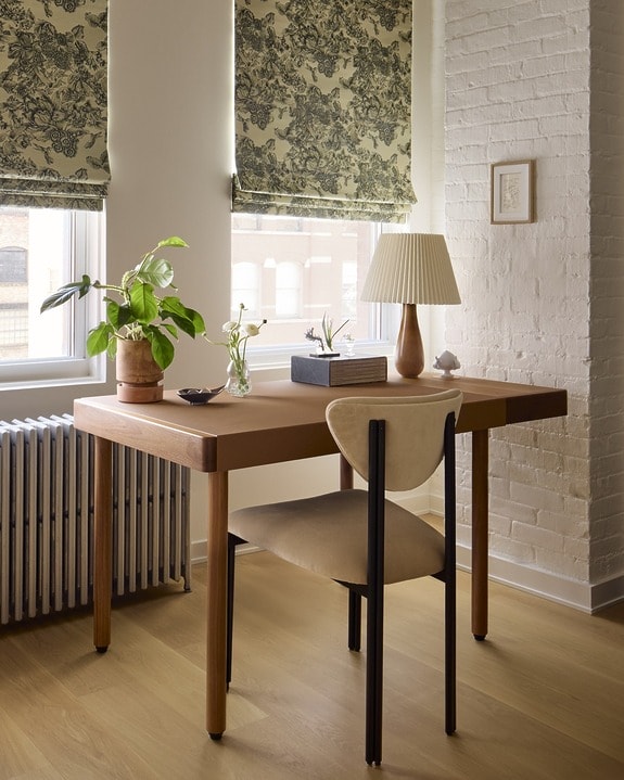 Cozy home office with a wooden desk, potted plants, books, and a lamp near windows with patterned blinds in natural light.