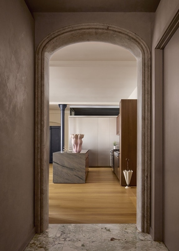 View through an arched doorway into a modern kitchen with wooden floors and a large decorative ceramic vase on the island.