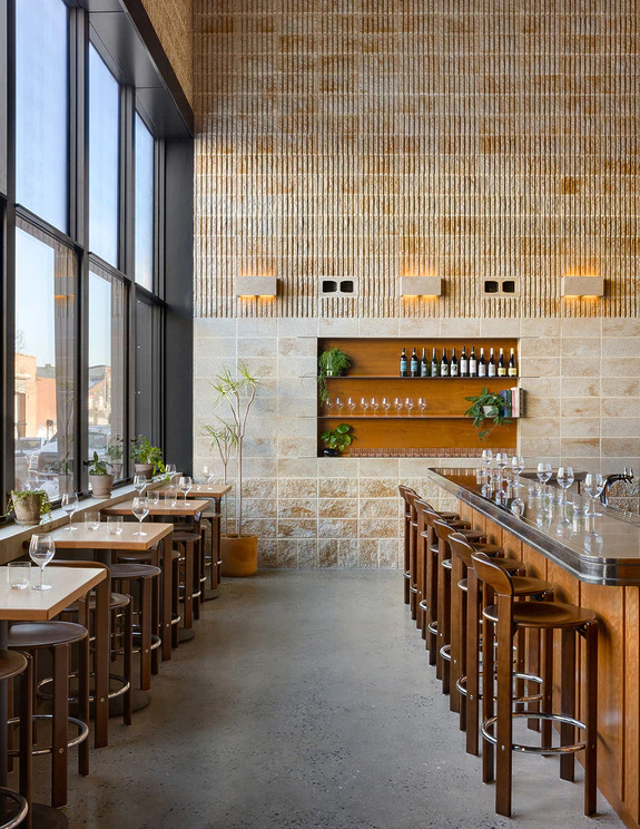 Modern, sunlit restaurant interior with wooden tables, tall windows, and shelves displaying wine bottles and plants.