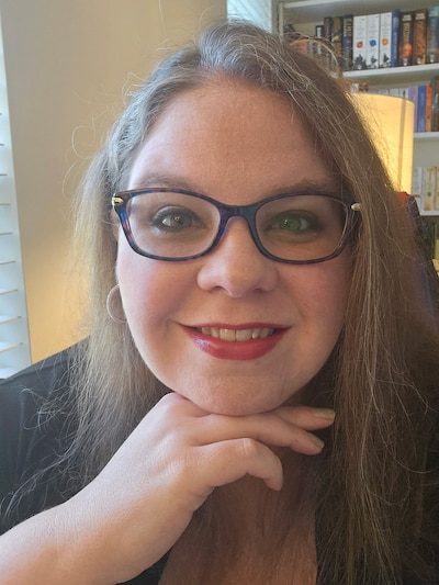 A white woman with long blonde hair wears dark rimmed glasses, smiles for a portrait in a house with books and a lamp in the background.