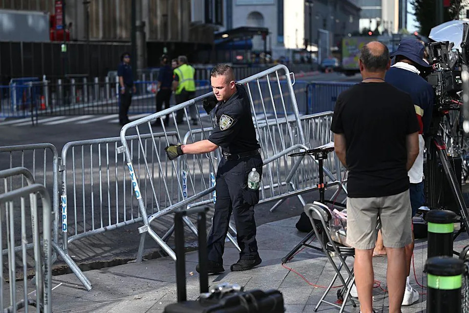 A police officer places a metal barricade on the streets around Madison Square Garden while a TV crew looks on.