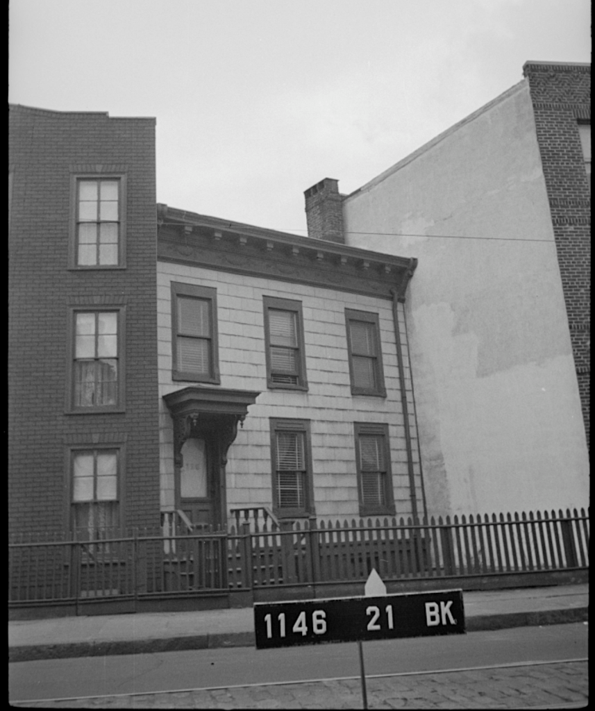 A black and white photograph of a historic building with a wooden porch, surrounded by brick structures and a fence. A street sign in the foreground displays the numbers '1146 21 BK'.