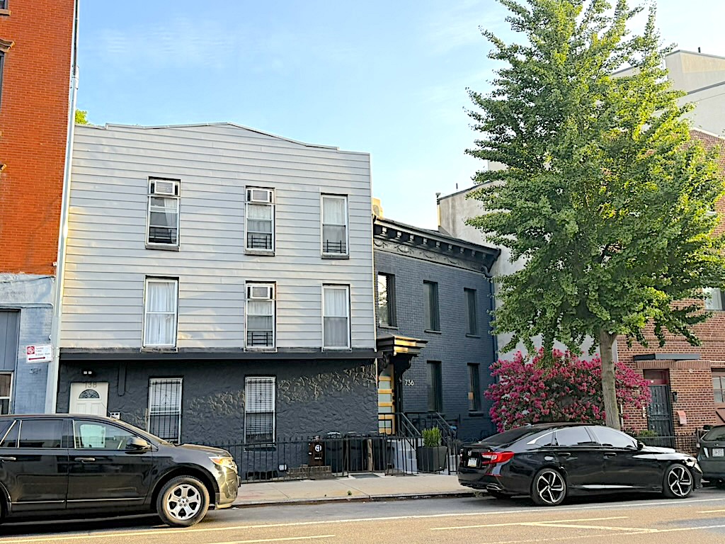 A street view of two neighboring buildings, one with a gray facade and multiple windows, and the other with a dark exterior and decorative elements. Black cars are parked along the roadside, and a green tree and flowering bushes add color to the scene.