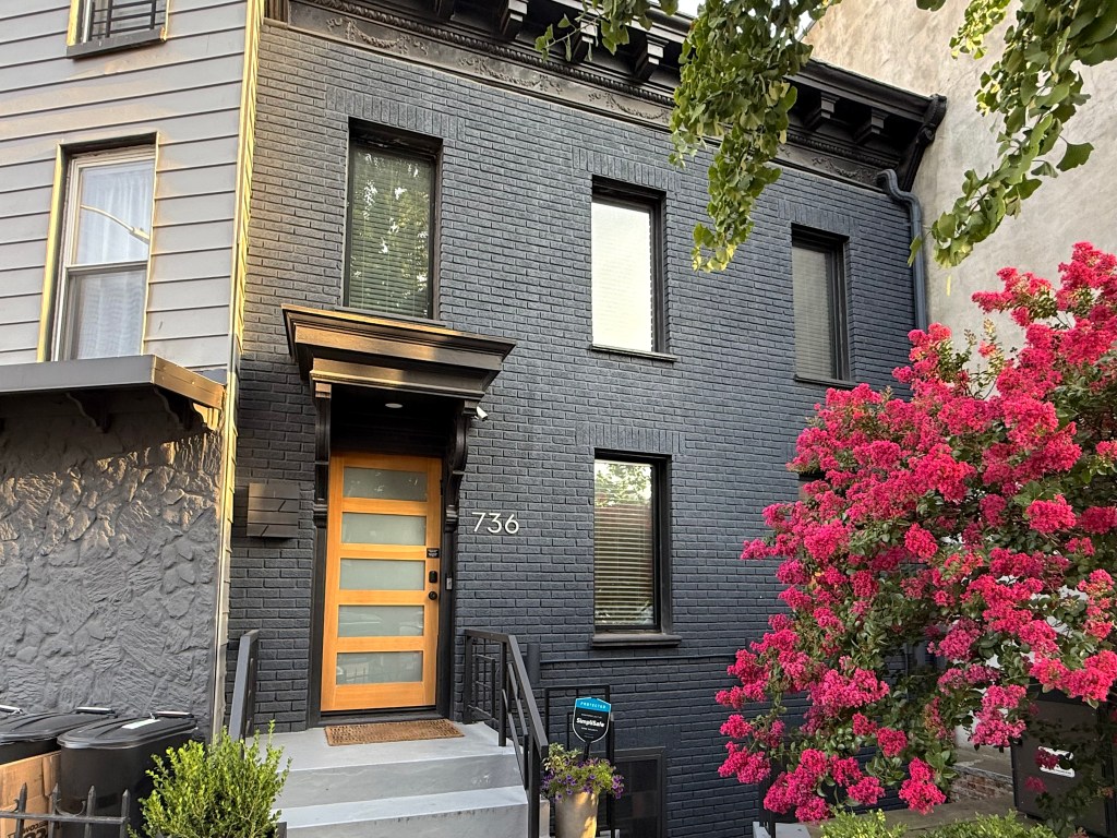 A modern two-story building with a black brick exterior and large windows, featuring a wooden front door with a glass panel. The entrance is framed by a small staircase and surrounded by vibrant pink flowering bushes.