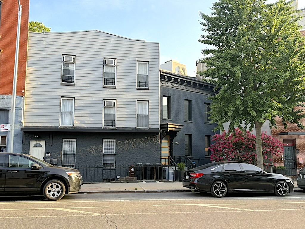 A street view showing two buildings: a gray multi-story building with multiple windows and an adjacent black building with distinct architectural features, surrounded by parked cars and greenery.