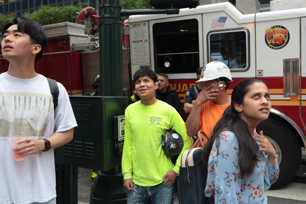 Workers, bystanders and firefighters gaze up at the Pfizer building in Midtown Manhattan after it was evacuated and at risk of collapse, July 7, 2026.