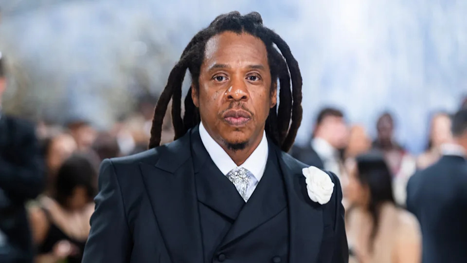 Jay-Z with long dreadlocks in a formal suit with a white flower pin, standing in a crowded event setting.