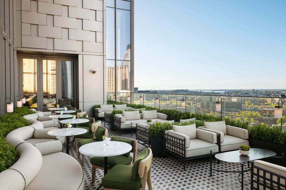 Stylish rooftop lounge area with comfortable seating, tables, greenery, and a panoramic city view in clear weather. Stylish rooftop lounge area with comfortable seating, tables, greenery, and a panoramic city view in clear weather.