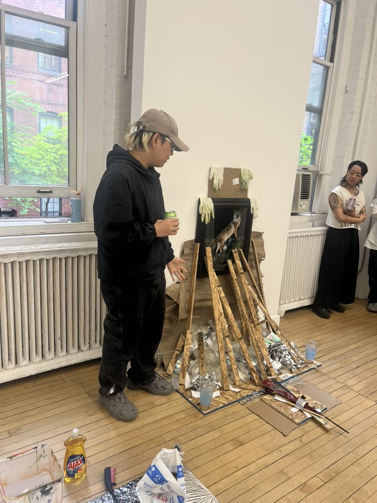 In an art studio, a young person in dark clothing and a cap stands beside an unusual installation made of wooden sticks and various materials. They are holding a drink and interacting with a group of observers, one of whom is standing to the right. The background features large windows allowing natural light, and the floor has paint supplies scattered around, including a container of dish soap.