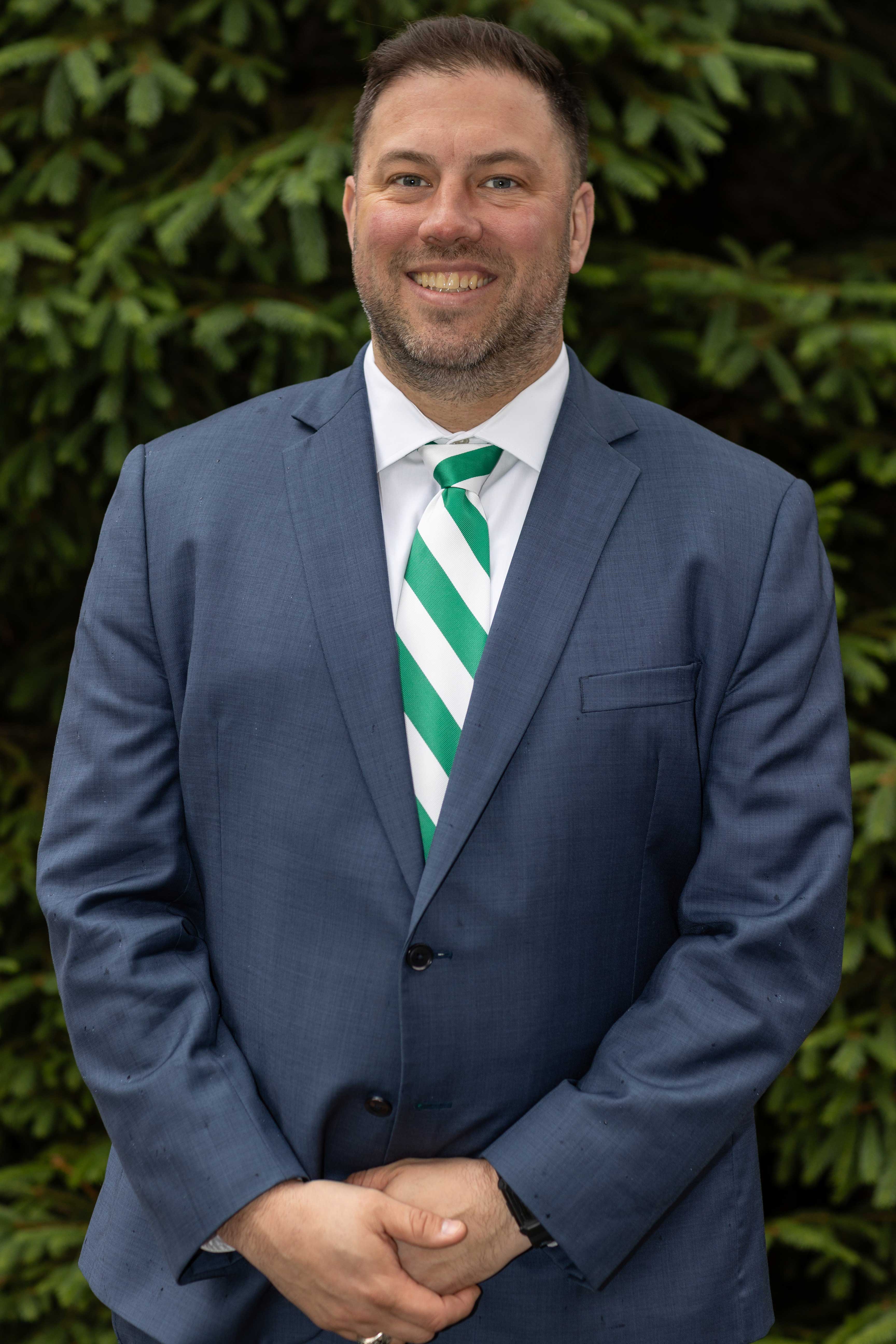 Professional portrait of a Ben Boivin in a gray suit and green and white diagnonally striped tie with lush green foliage in the background.