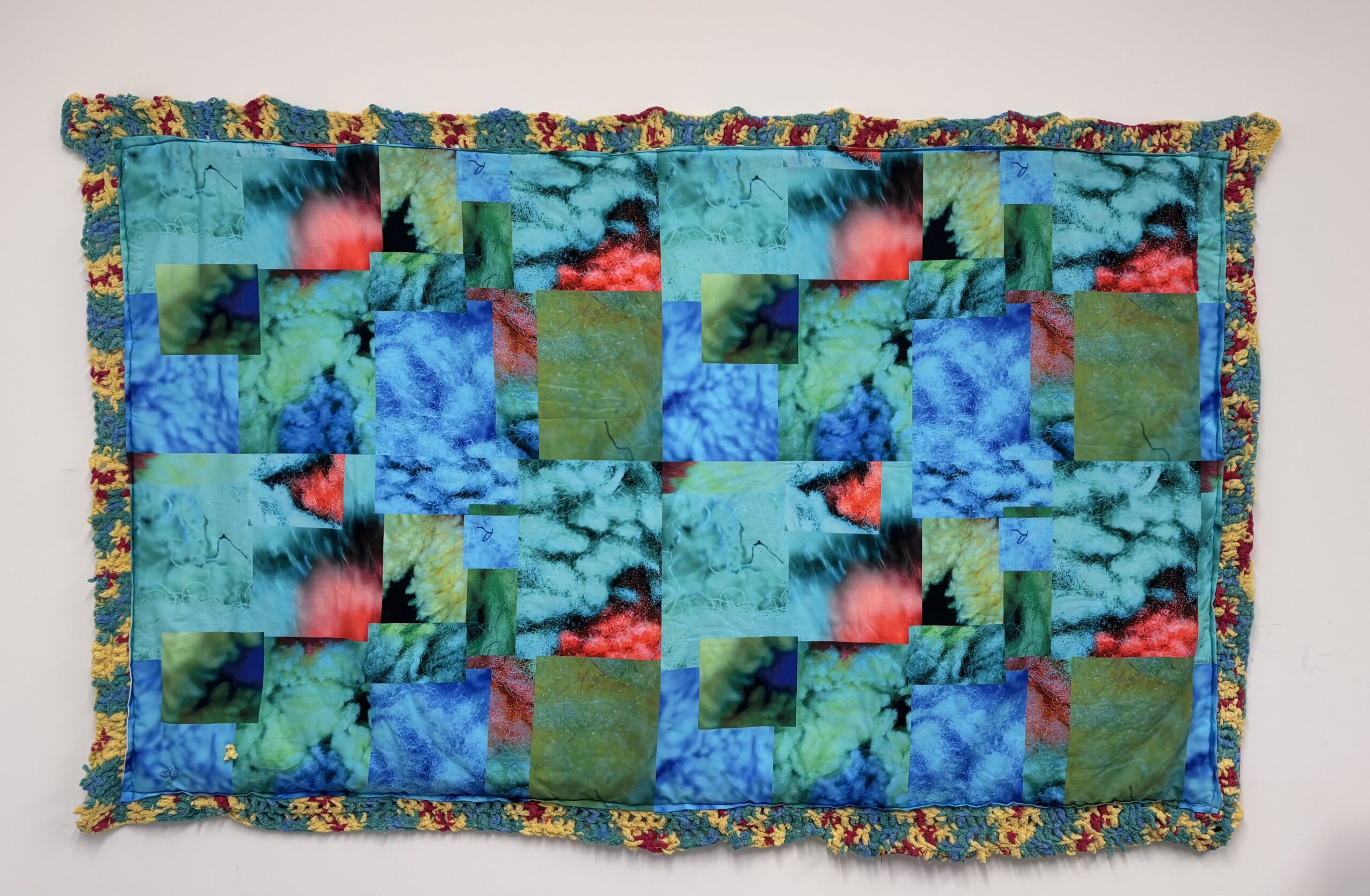 A colorful quilt displayed on a white wall. The quilt features an abstract pattern with overlapping squares in vibrant shades of blue, green, red, and yellow. The edges are bordered with a textured trim that includes green, yellow, and red yarn, adding a decorative touch to the overall design.