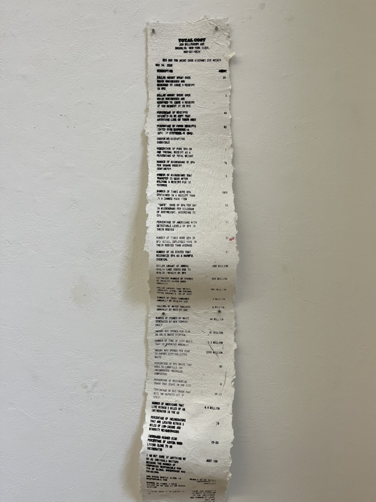 A long, vertical receipt printed on textured paper hangs against a white wall. The receipt includes various charges, fees, and totals, presented in a structured format. The top section states "TOTAL COST" and lists items with accompanying costs, including several large figures. It also contains references to additional services or fees. The overall appearance is slightly worn, with frayed edges.