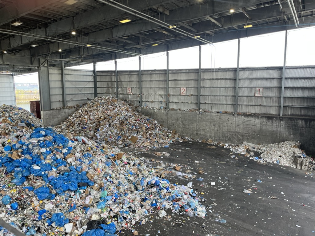 An indoor waste management facility with large piles of mixed waste material, including cardboard, plastic wrappers, and other garbage. The structure has high ceilings and is well-lit, with metal walls and a concrete floor. There are numbered markers on the walls, indicating sections within the facility. Natural light enters through the large windows.