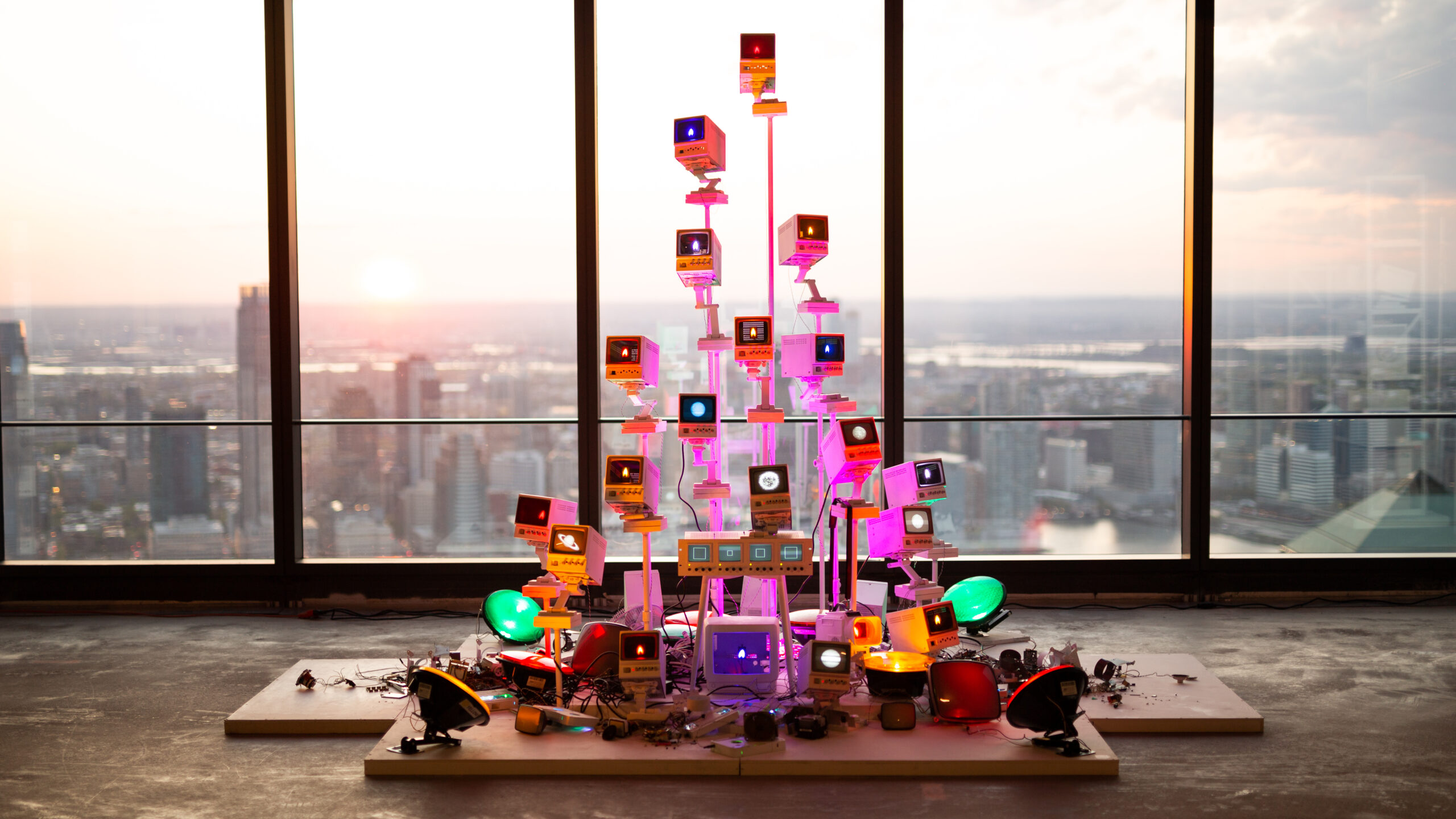 A colorful installation made of stacked vintage monitors and lights, set against large windows with a city skyline in the background during sunset. The monitors display various glowing colors, surrounded by other electronic components on a wooden platform. The warm hues of sunset reflect off the glass, enhancing the vibrant atmosphere of the artwork.