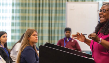 Female instructor standing at a classroom podium addressing a group of teenage students.