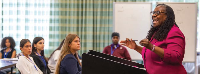 Female instructor standing at a classroom podium addressing a group of teenage students.