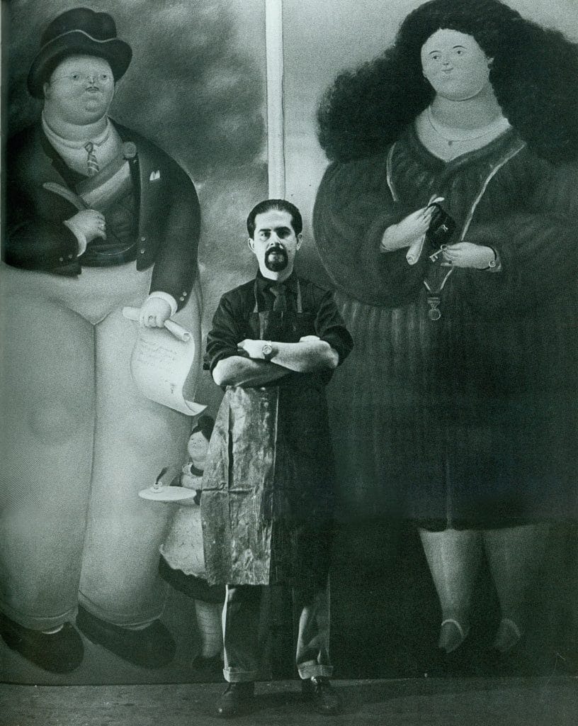 Fernando Botero stands between oversized portraits exemplifying his signature voluminous figures during an early studio photograph.