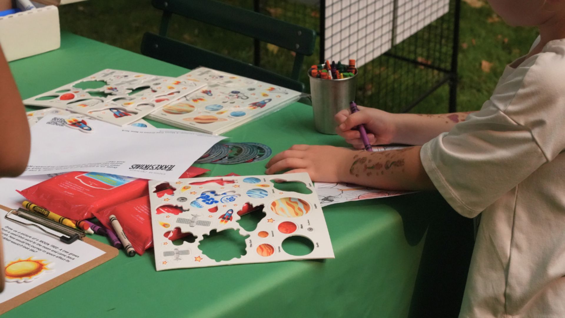 A young boy drawing with crayons also has temporary space tattoos on his arm.