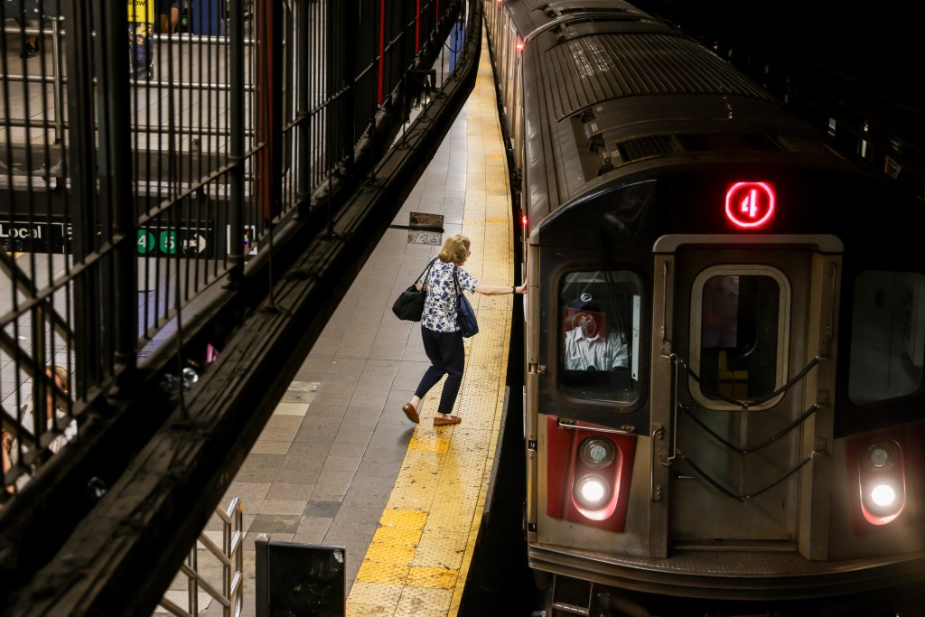 A woman in a floral shirt and dark pants stands on a subway platform next to a stopped train.