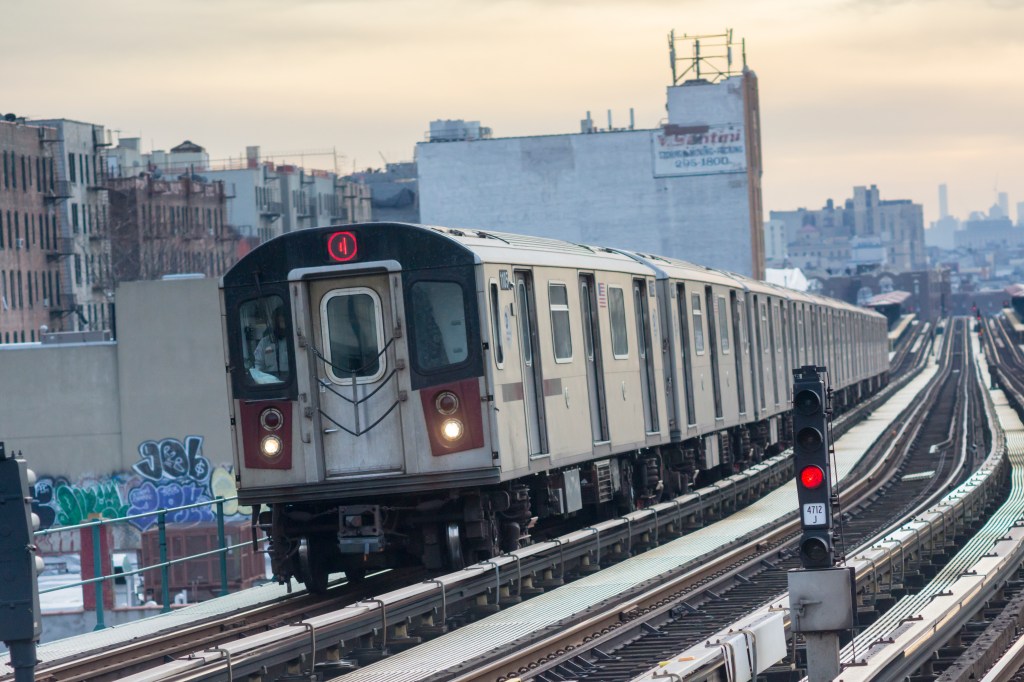 A subway train with a red "1" on the front travels on elevated tracks in a city with buildings in the background.
