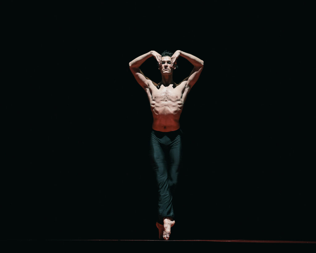 Onstage in a spotlight, Hugo Marchand stands in sous-sus with his back foot on releve and his front pointed straight down. He lifts his arms, elbows and wrists bent, so that his fingertips frame the sides of his eyes. He wears black pants and has a serious expression.