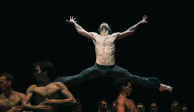 Hugo Marchand, surrounded by an ensemble of dancers, jumps with his legs split sideways and his arms stretched out to the sides. He tilts his head back and splays his fingers. He and the other dancers all wear black pants.