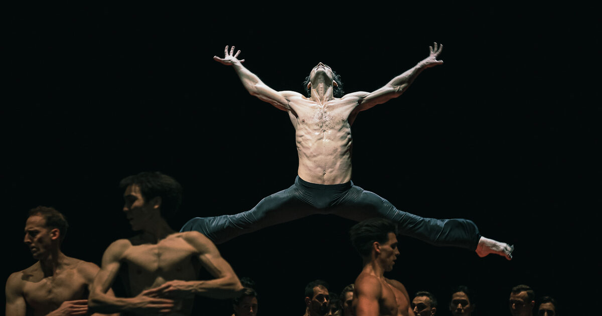 Hugo Marchand, surrounded by an ensemble of dancers, jumps with his legs split sideways and his arms stretched out to the sides. He tilts his head back and splays his fingers. He and the other dancers all wear black pants.