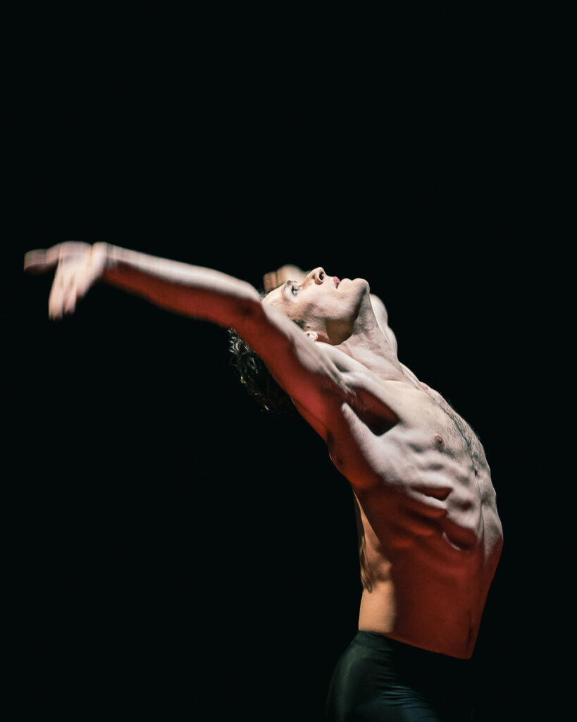 Hugo Marchand, shown from the hips-up in a spotlight, dances onstage wearing black pants. He arches up and back, his arms wide with his palms up and fingers down, lips slightly parted as he looks up.