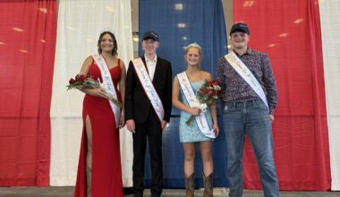 Four pageant contestants stand on a stage in front of red, white, and blue curtains, wearing sashes and crowns, and holding bouquets.