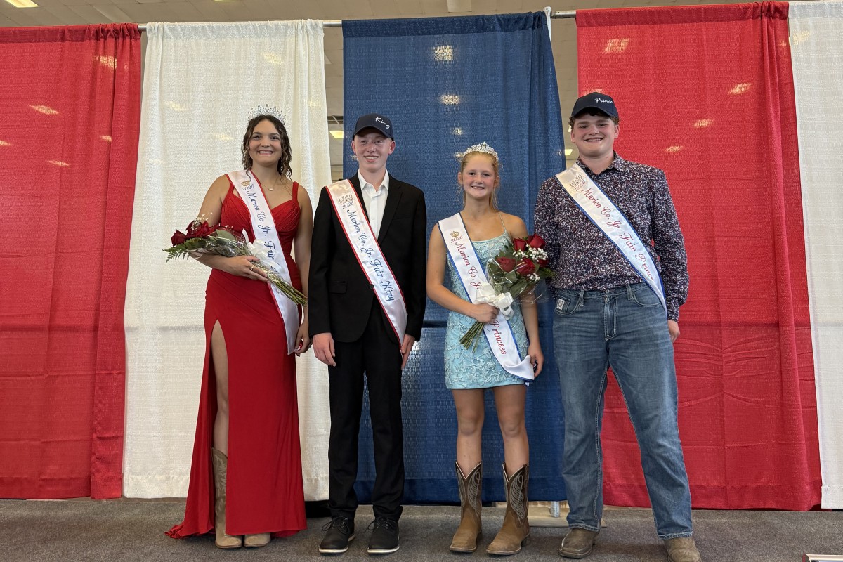 Four pageant contestants stand on a stage in front of red, white, and blue curtains, wearing sashes and crowns, and holding bouquets.