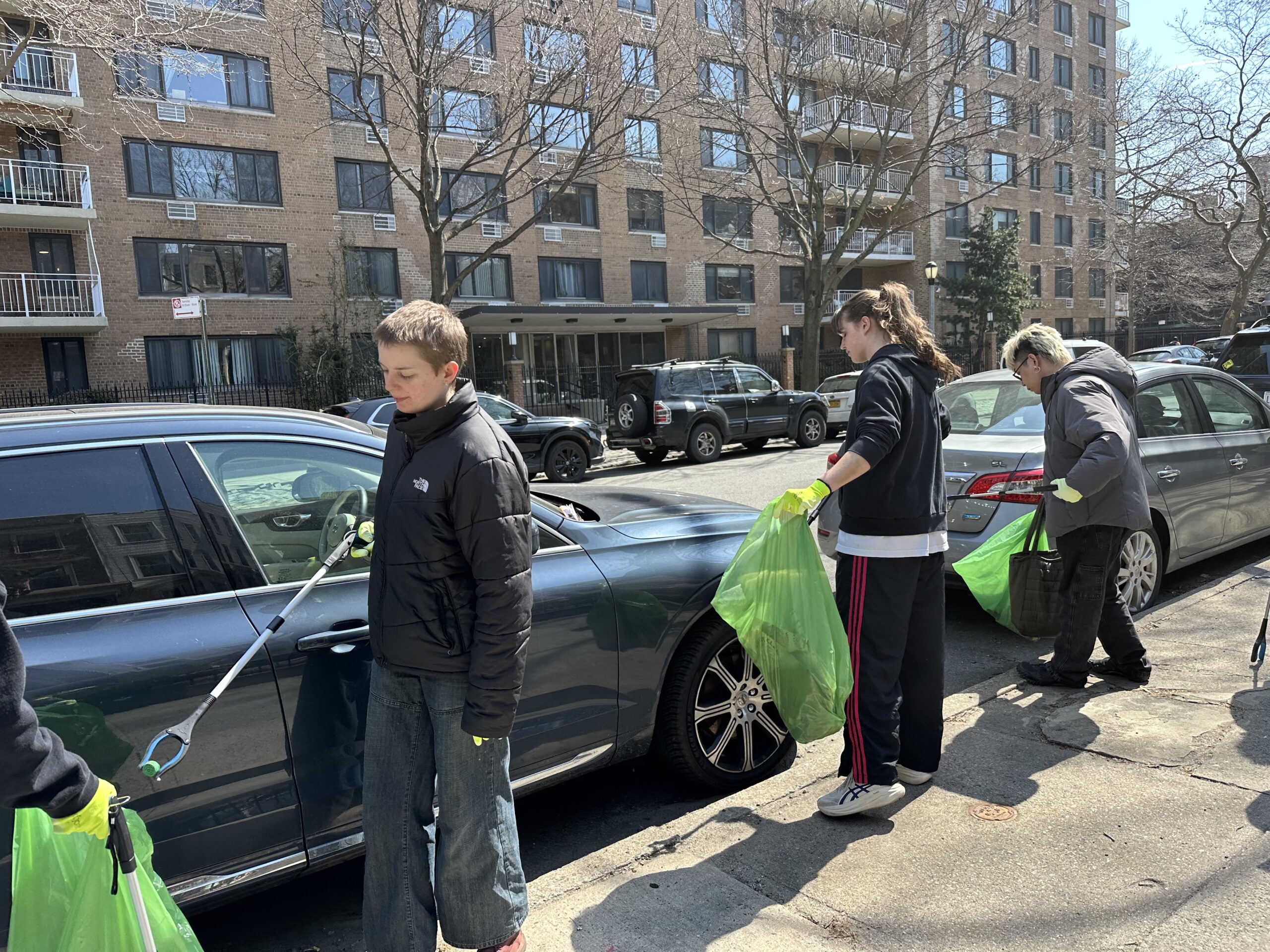 A group of four individuals is participating in a cleanup activity on a city street. They are wearing gloves and holding green trash bags. One person uses a grabber tool to pick up debris near a parked car, while others carry bags filled with collected waste. In the background, several residential buildings and parked vehicles are visible, indicating an urban environment on a sunny day.