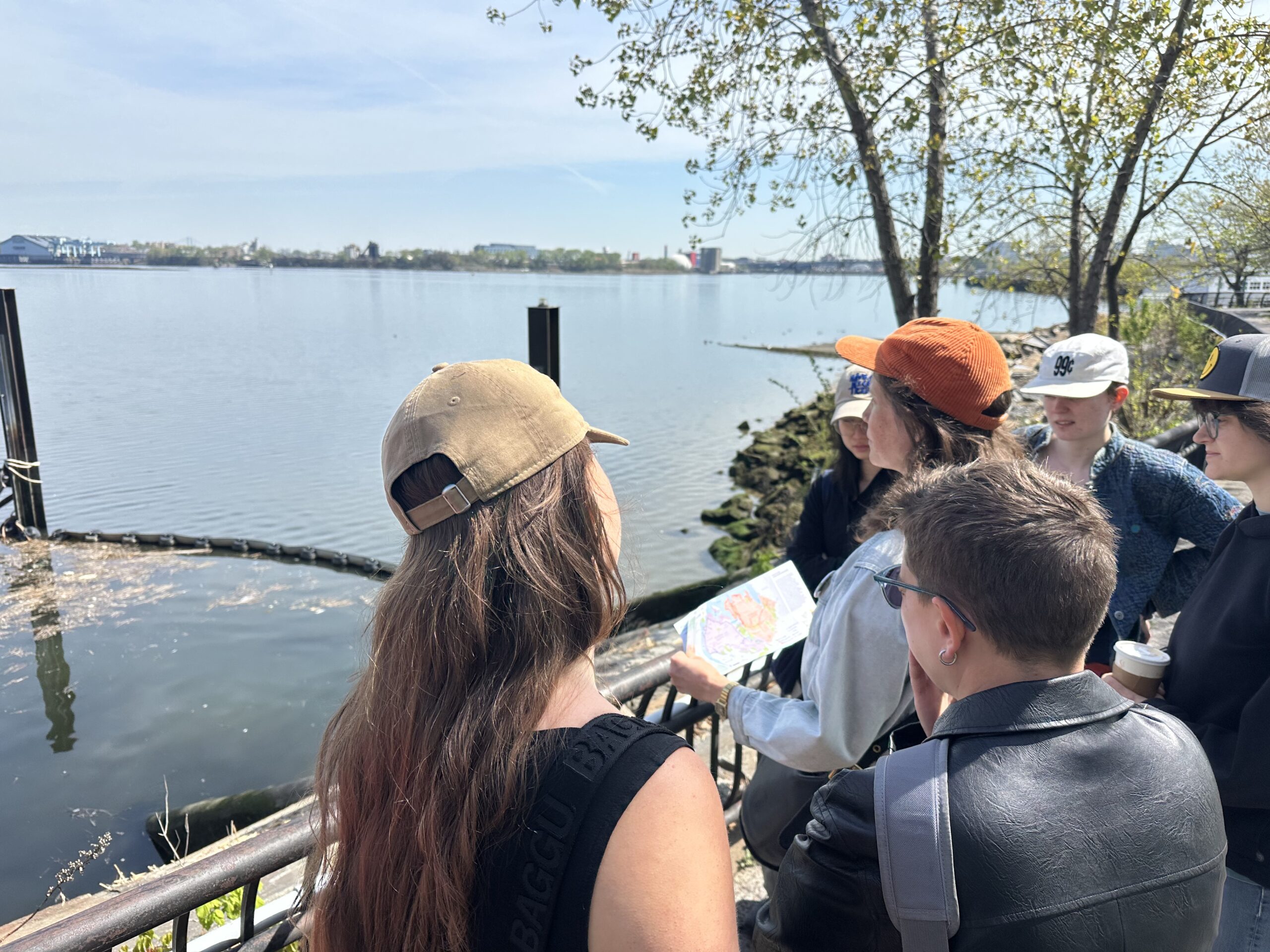 A group of people stands near a body of water, looking towards the shore. One person holding a map is engaged in discussion, while others listen closely. The background features trees with budding leaves and distant buildings. The sky is clear and sunny, reflecting a pleasant day.
