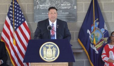 Assemblyman Brian Manktelow speaks at a podium with the New York State seal, flanked by American and New York flags.
