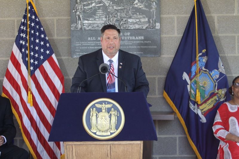 Assemblyman Brian Manktelow speaks at a podium with the New York State seal, flanked by American and New York flags.