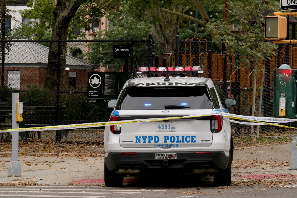 A white NYPD Police car with flashing red and blue lights, with yellow crime scene tape across the back of the vehicle, in front of a playground.