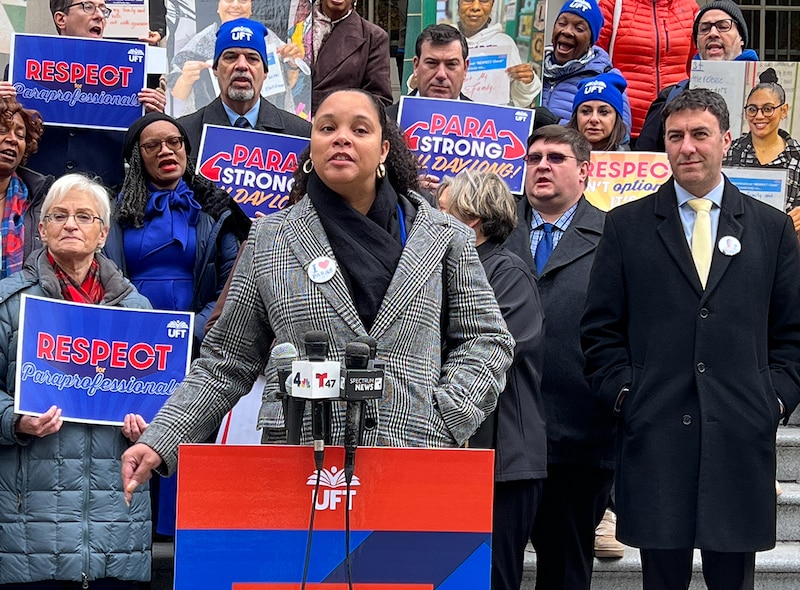 A woman in a gray plaid coat and black sweater speaks at a podium in front of people holding signs.