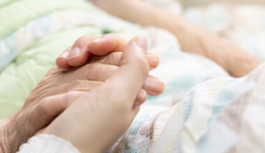 A close-up of two people holding hands, one appearing elderly and the other younger, resting on a bed with a green blanket and patterned sheets, conveying comfort and support.