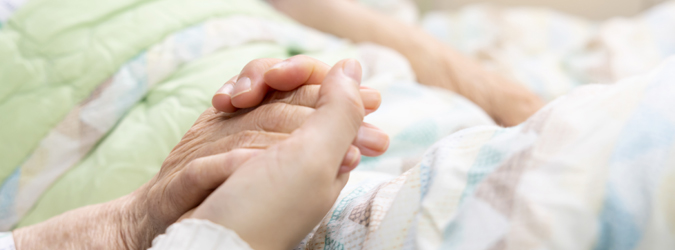 A close-up of two people holding hands, one appearing elderly and the other younger, resting on a bed with a green blanket and patterned sheets, conveying comfort and support.