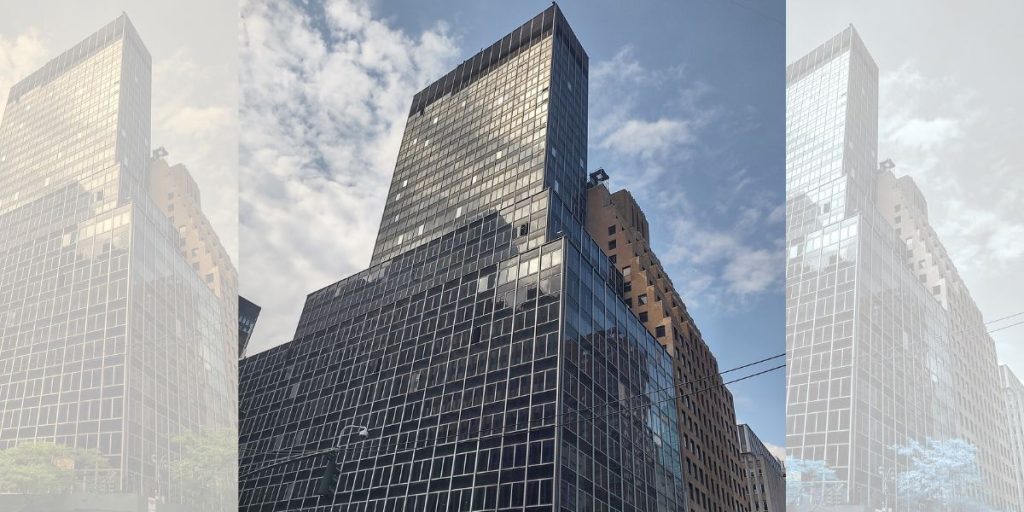 A towering glass and steel skyscraper against a partly cloudy blue sky. The building reflects sunlight, creating a sleek and modern appearance.