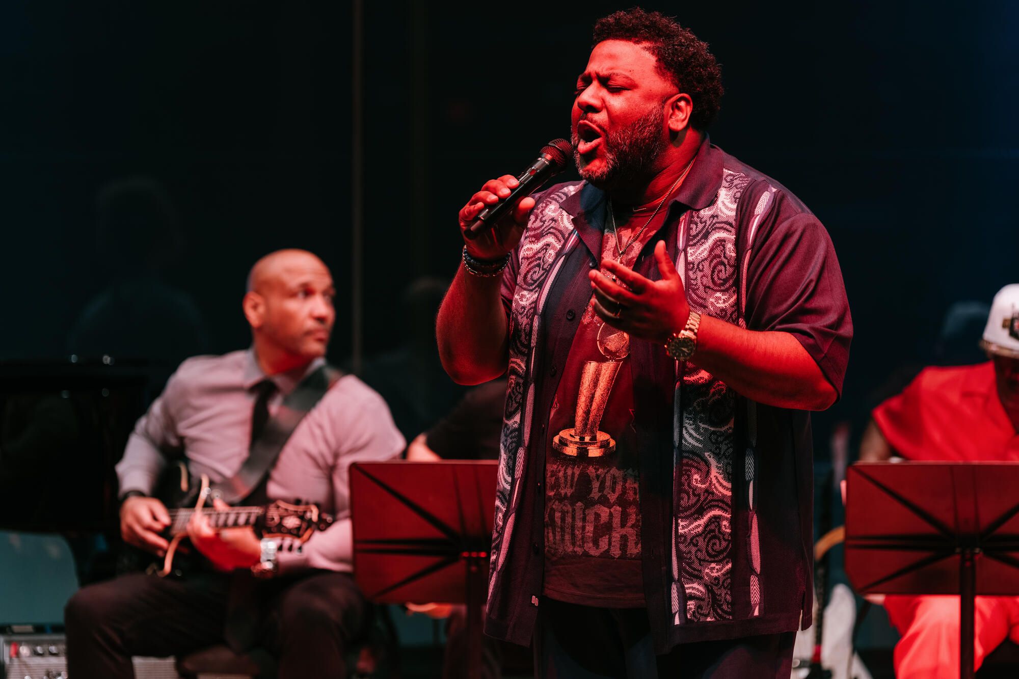 A photo shows a Black man wearing a patterned short-sleeved button-down shirt over a T-shirt while singing into a microphone. To the left is a man with brown skin playing a guitar while sitting down. 